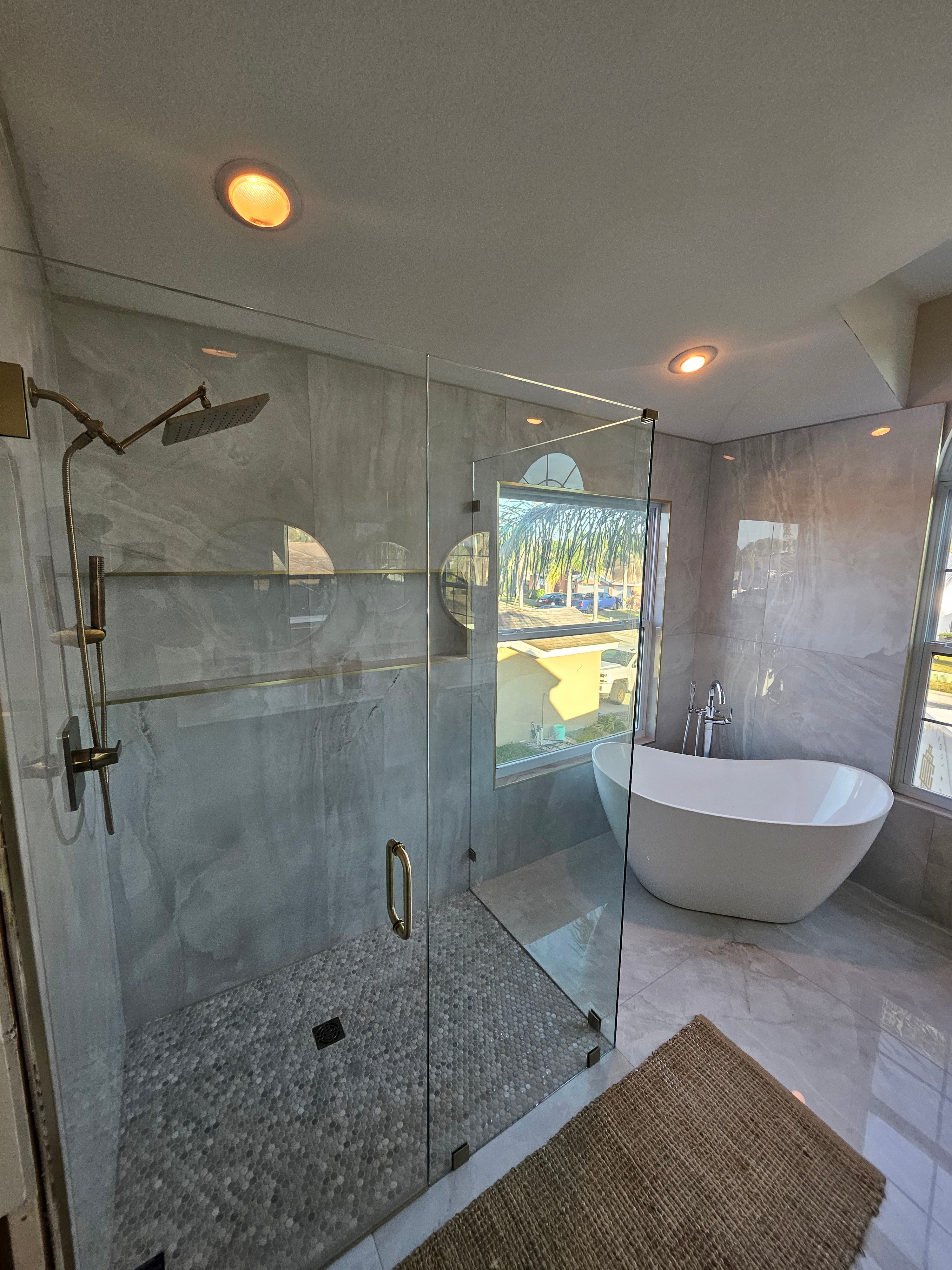 Finished Tampa Bay master bathroom with a frameless glass walk-in shower, large-format marble-look porcelain tile, pebble mosaic shower floor, brushed-gold fixtures, and a freestanding soaking tub
