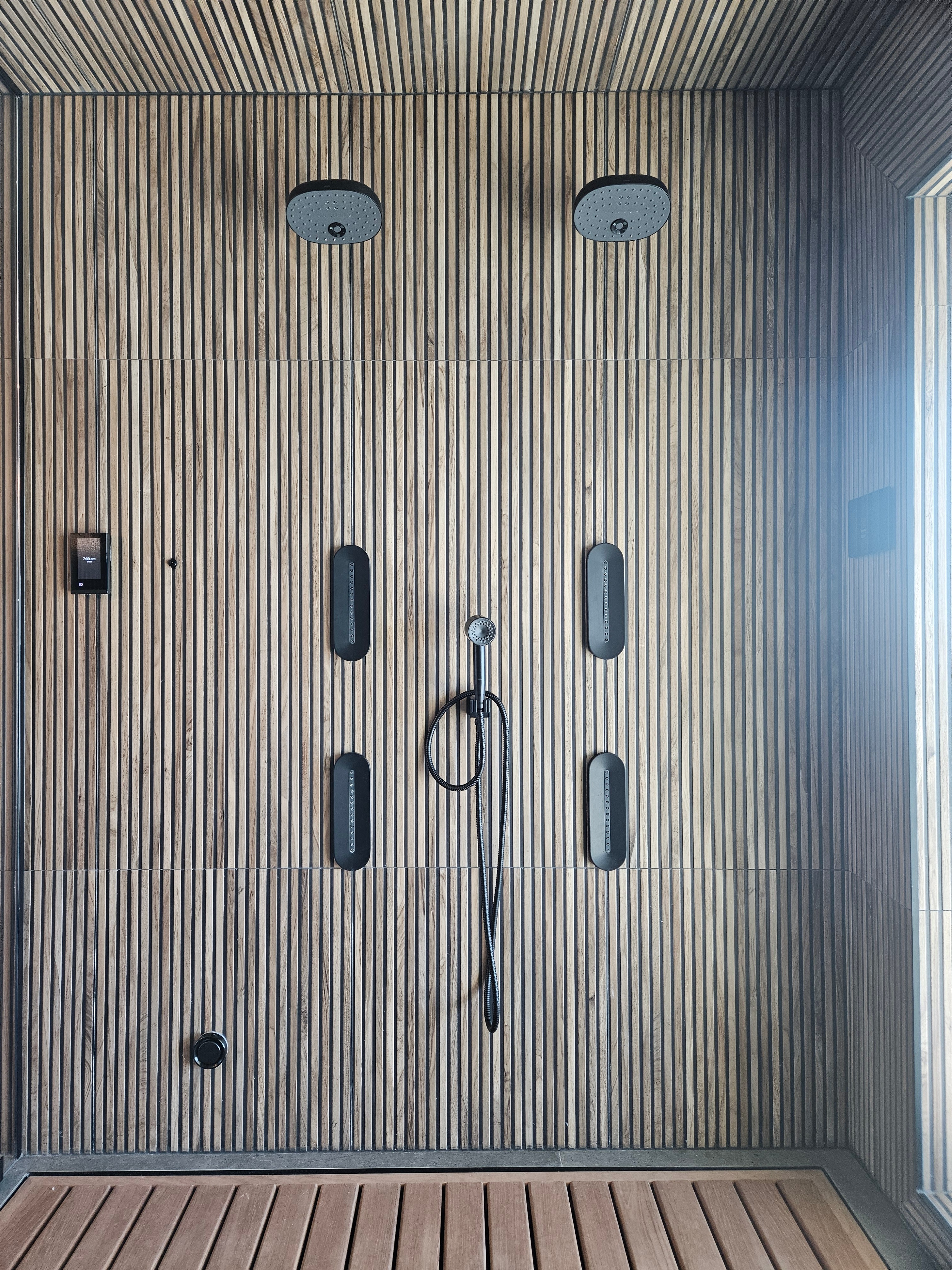 Luxury wood-slat shower with two matte-black ceiling rain heads, four wall body-spray jets, a handshower, and a digital touchscreen control all installed together