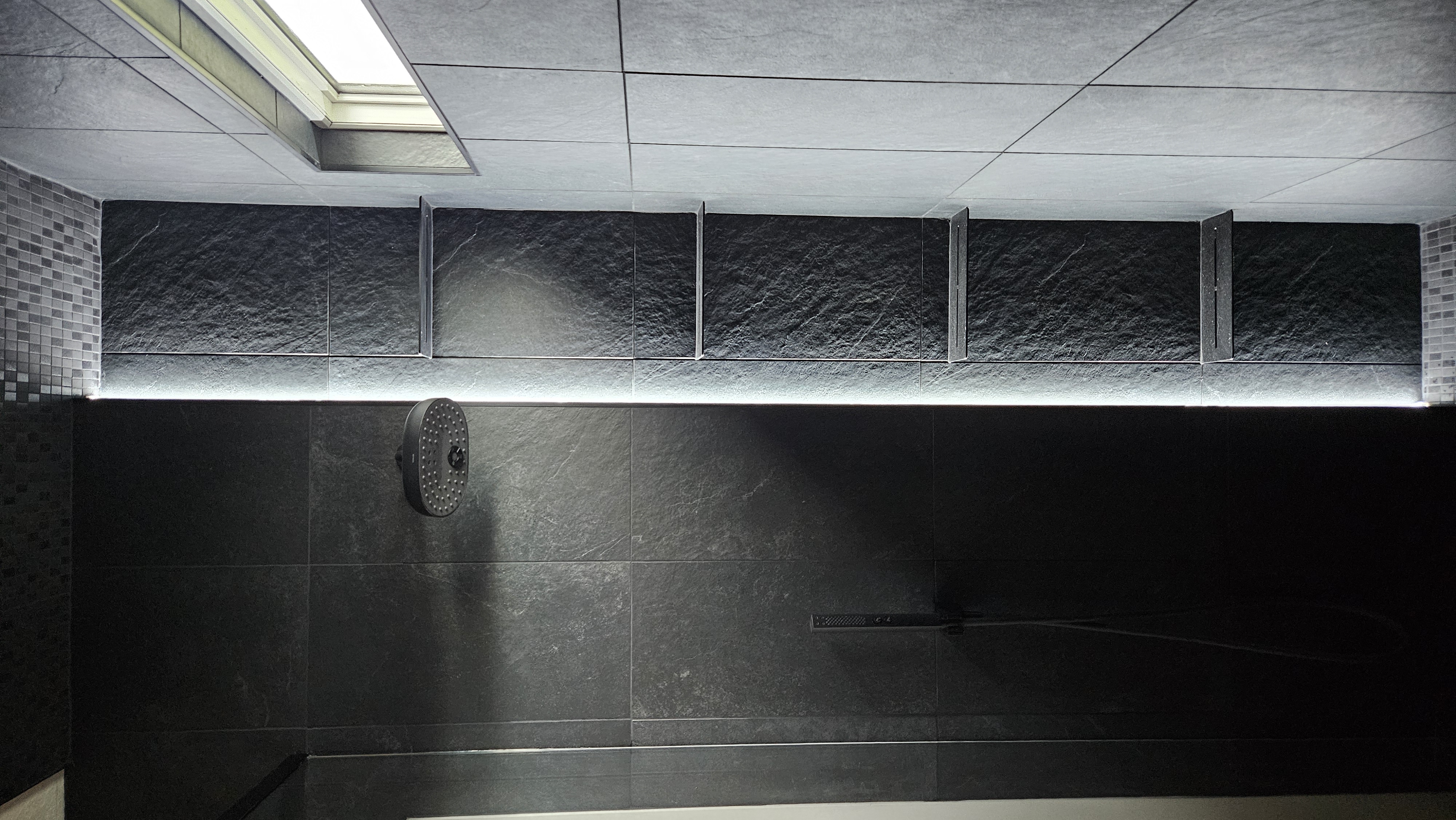 Matte-black rain showerhead and handshower beside a column of floating slate niche shelves